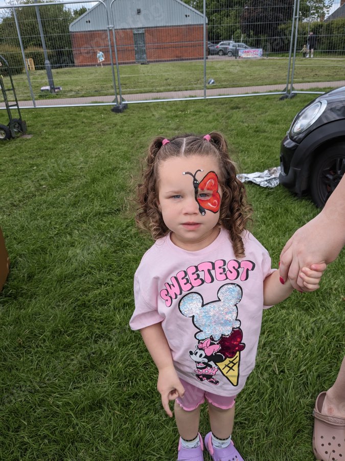 Toddler with a red butterfly cheek painting at an outdoor event