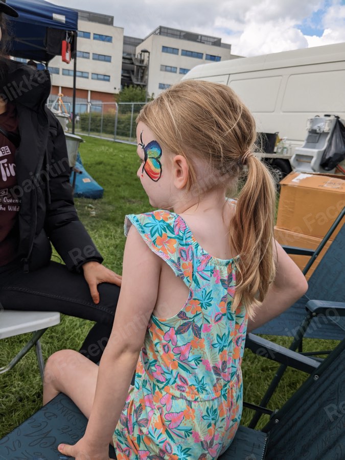 A child at Relikka's face painting stall at an outdoor fun day, showing a rainbow butterfly cheek design